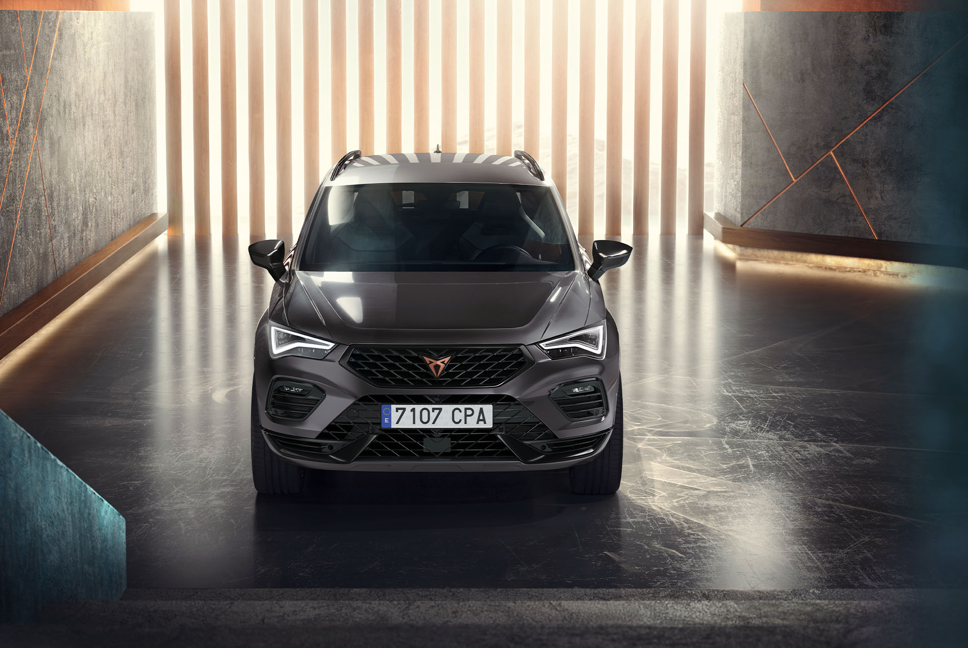  Vista frontale del SUV Cupra Ateca, colore grigio, in un ambiente moderno e illuminato. L'immagine mette in evidenza l'audace griglia frontale della vettura, i gruppi ottici a LED e il design elegante. 