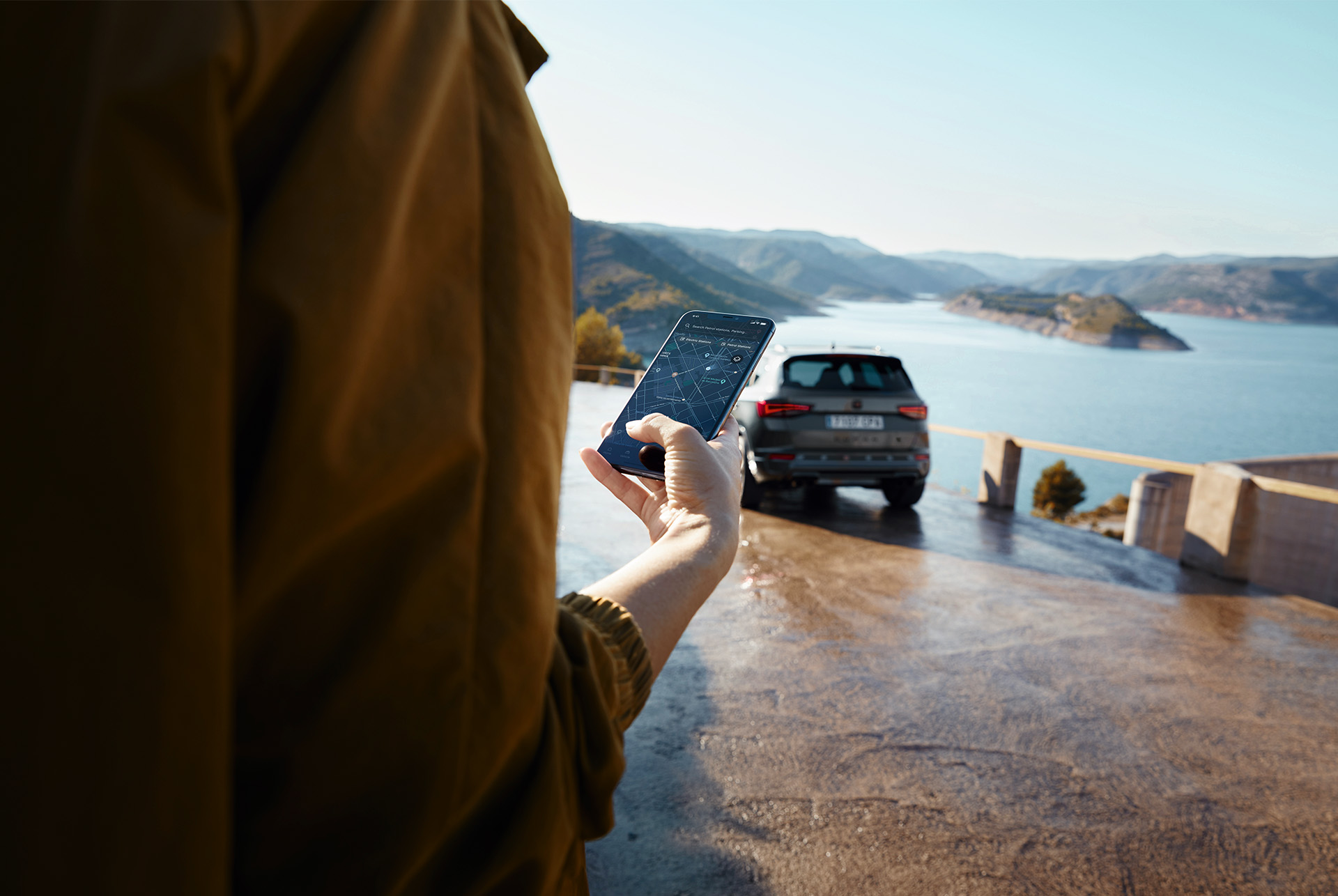 Una persona mentre tiene in mano uno smartphone con CUPRA Ateca sullo sfondo, parcheggiata vicino a una vista panoramica sulle montagne e sull'acqua, simbolo di un mondo connesso.