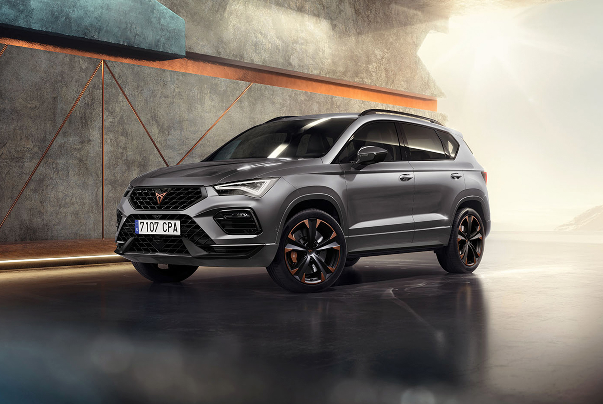 Il suv sportivo e compatto cupra ateca 2024 in graphite grey parcheggiato in un ambiente moderno e industriale. Il design della vettura è caratterizzato da linee aerodinamiche, gruppi ottici a LED ed esclusivi cerchi in lega con finiture in rame. Lo sfondo include pareti dli motivi geometrici, in contrasto con l'elegante design del veicolo.