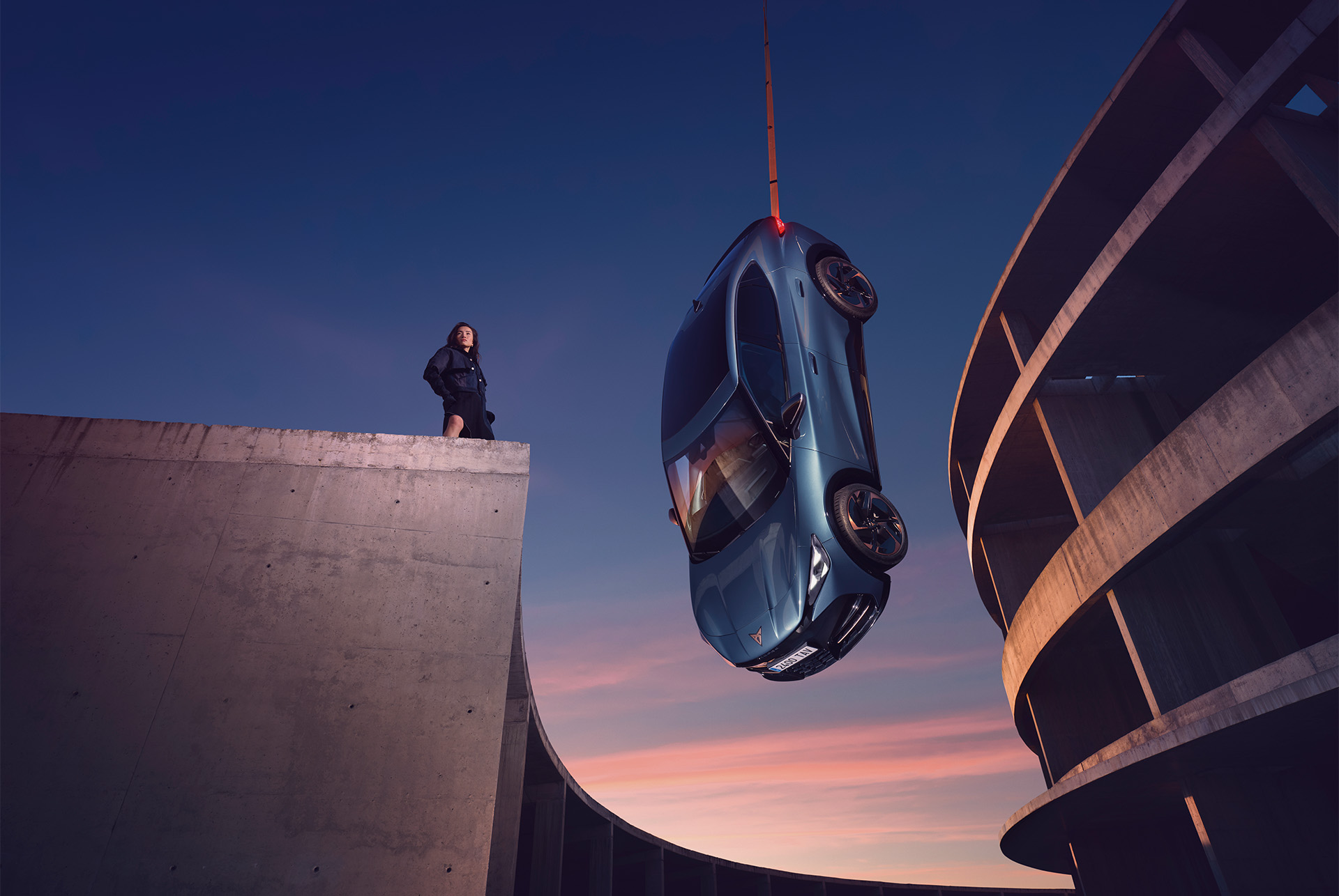 SUV elettrico CUPRA TAVASCAN 2024 blu con cerchi in lega color rame e fari triangolari, sospeso a mezz'aria sullo sfondo dell'architettura in cemento durante il tramonto